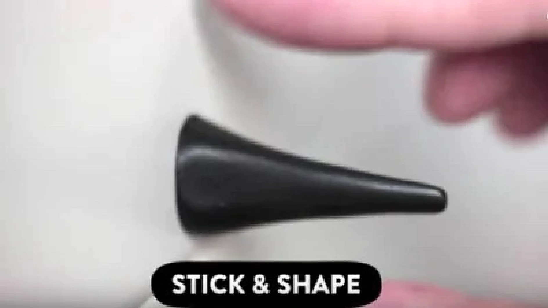Die Hand einer Person formt ein kleines, schwarzes, kegelförmiges Objekt vor einer weißen Oberfläche. Die Worte „STICK & SHAPE“ werden in einem schwarzen Kästchen am unteren Bildrand angezeigt, was die Verwendung von Tesa-Klebeband verdeutlicht. (This text has been generated by AI)