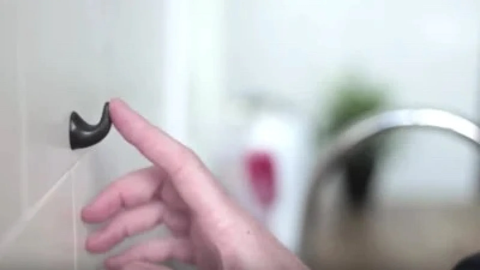Nahaufnahme einer Hand, die mit einem kleinen, gebogenen schwarzen Haken interagiert, der mit Tesa-Klebeband an einer Wand befestigt ist. Im Hintergrund sind ein verschwommener Seifenspender und eine grüne Pflanze zu sehen. Die Szene scheint in einem Badezimmer oder einer Küche zu spielen. (This text has been generated by AI)