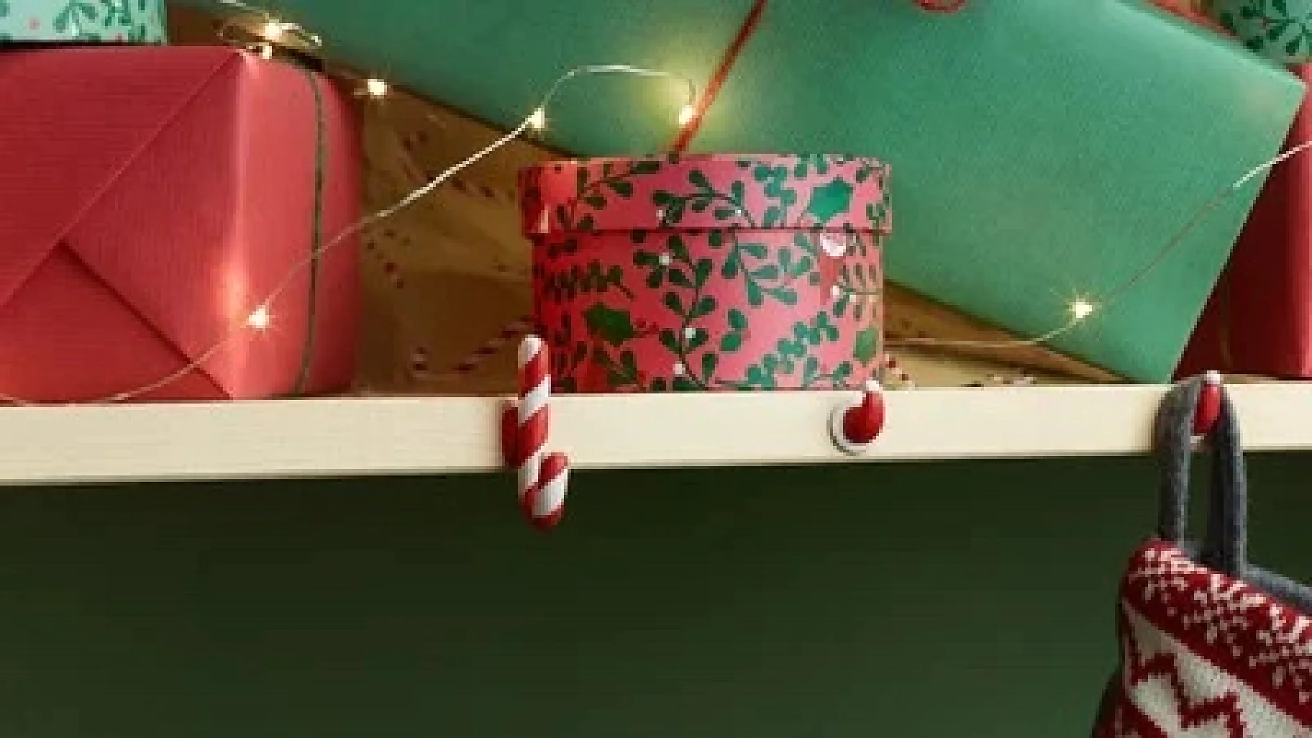 Ein Regal, geschmückt mit eingepackten Geschenken und Lichterketten. Auf dem Regal steht eine zylindrische Geschenkbox mit einem rot-grünen Stechpalmenmuster. Darunter hängen Haken für Zuckerstangen, von denen einer etwas hält, das wie ein Strumpf aussieht. (This text has been generated by AI)