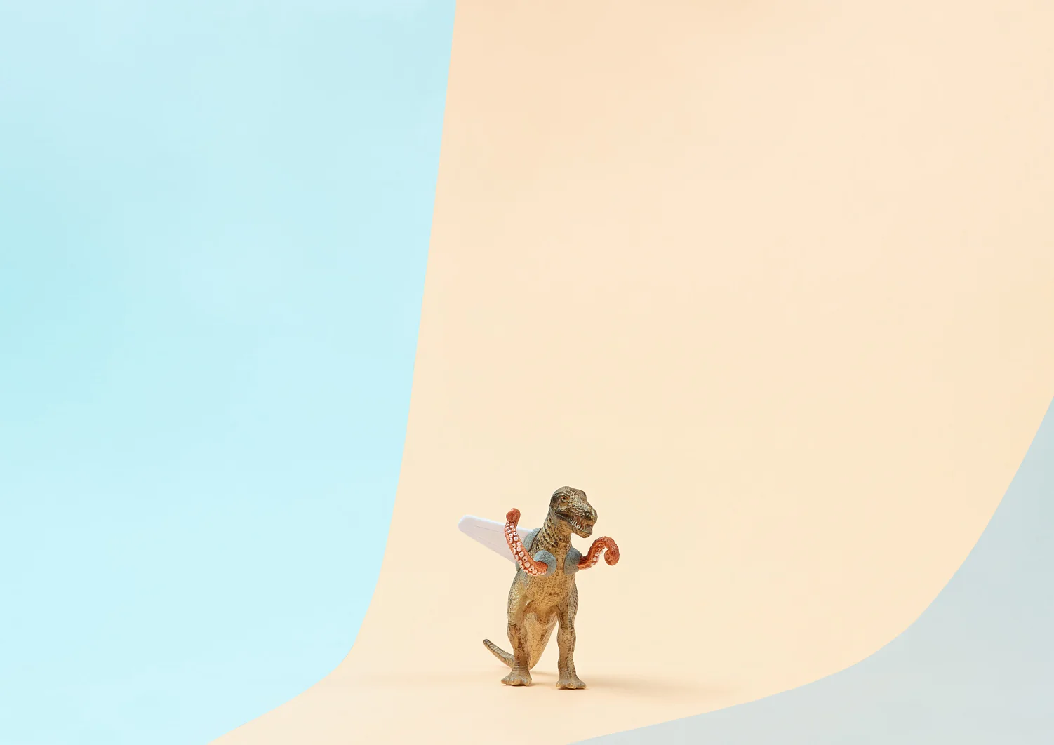 Ein Spielzeugdinosaurier steht auf einer sanft geschwungenen Oberfläche mit blauem Hintergrund auf der linken und beigem Hintergrund auf der rechten Seite. Der Dinosaurier hält einen kleinen Gegenstand, der einem Stück Kuchen ähnelt und mit Tesa-Klebeband befestigt ist. (This text has been generated by AI)