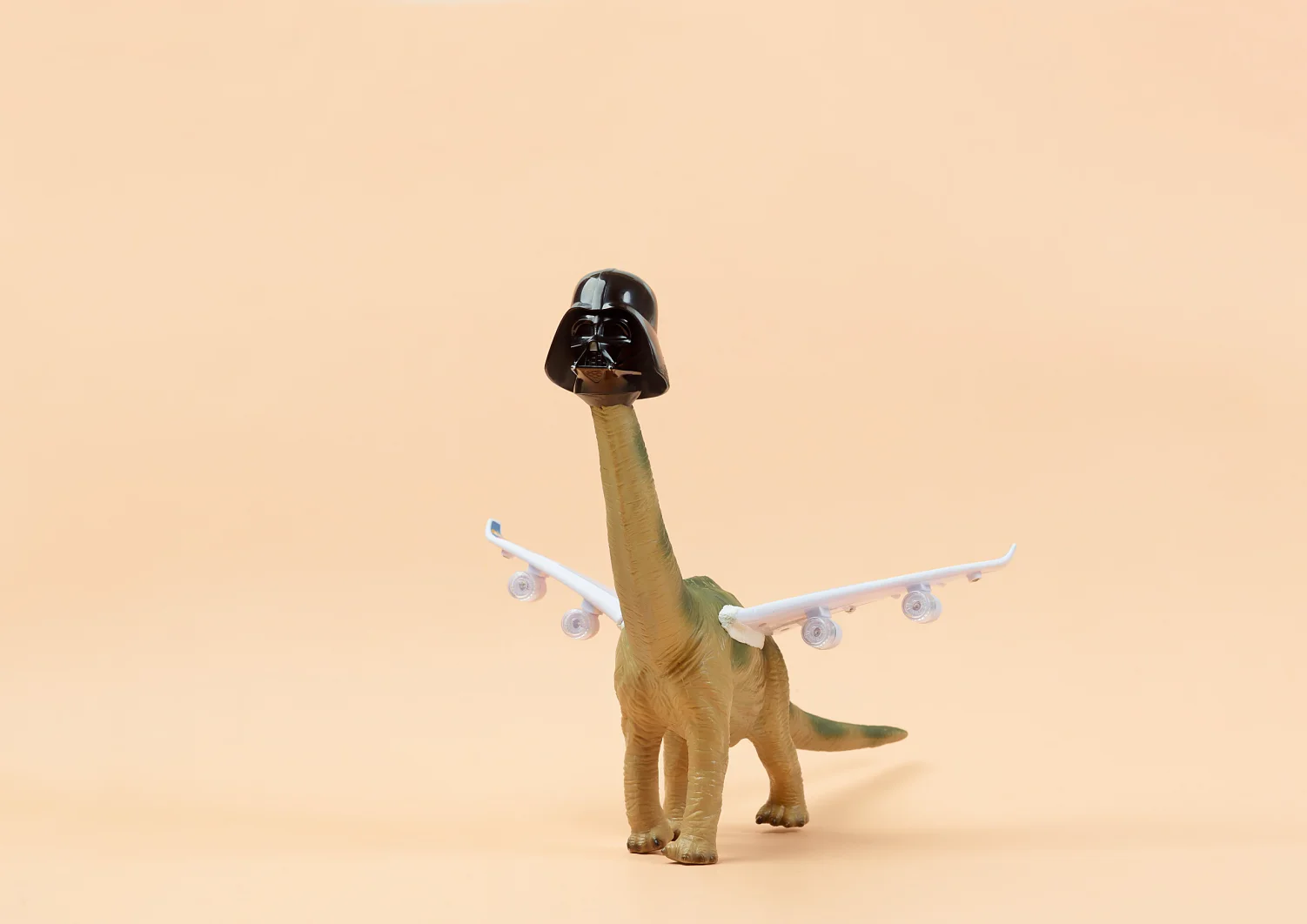 Ein Spielzeugdinosaurier mit langem Hals trägt einen schwarzen Helm, der an einen aus einem Science-Fiction-Film erinnert. An seinem Rücken sind Flugzeugflügel mit mehreren Triebwerken befestigt, die mit Tesa-Klebeband befestigt sind. Der Hintergrund ist in einem schlichten hellen Beige gehalten. (This text has been generated by AI)