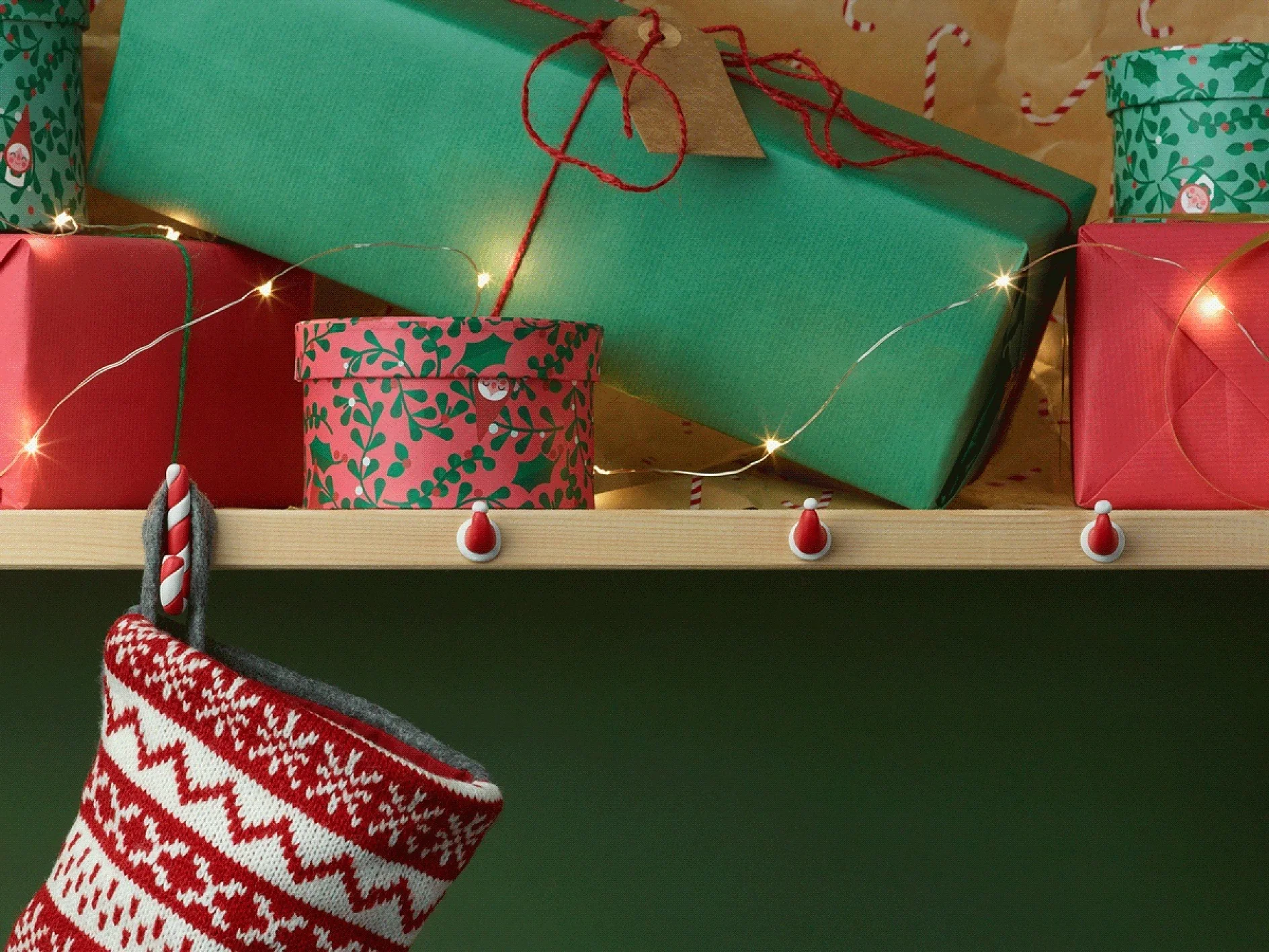 Auf einem Regal sind grün und rot verpackte Geschenke mit rot-weißem gemustertem Papier ausgestellt, alles mit Tesa-Klebeband befestigt. Eine Lichterkette schmückt das Regal. Darunter hängt ein rot-weiß gestrickter Weihnachtsstrumpf mit festlichem Muster. (This text has been generated by AI)
