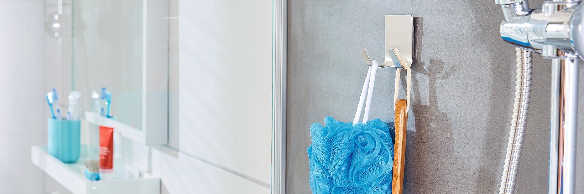 In einem Badezimmer mit grau gefliesten Wänden und einer Dusche hängt an einem Metallhaken ein blauer Badeschwamm aus Netzgewebe und eine Badebürste aus Holz. Rechts ist ein Duschkopf zu sehen. Im Hintergrund ist ein weißes Waschbecken mit Toilettenartikeln zu sehen, die mit Tesa-Klebeband befestigt sind. (This text has been generated by AI)