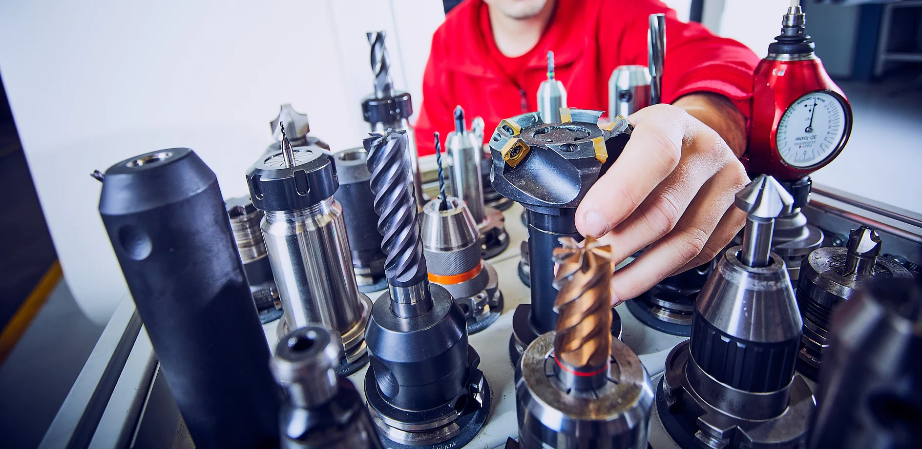 Eine Person in einer roten Jacke wählt ein Werkzeug aus einer Sammlung verschiedener Bohrer und Industriewerkzeuge aus, die auf einem Tisch in einer Werkstatt angeordnet sind. Die Werkzeuge sind aus Metall und variieren in Größe und Form. (This text has been generated by AI)