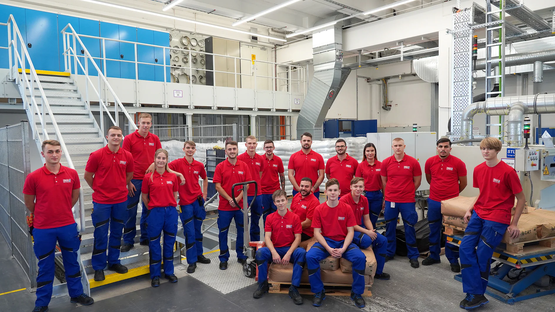 Eine Gruppe von 18 Personen in roten Hemden und blauen Hosen steht und sitzt auf braunen Säcken in einer Industrieumgebung. Sie stehen vor Maschinen und Metalltreppen, die in eine obere Etage führen. Der Bereich ist durch Deckenleuchten gut beleuchtet. Anstelle von herkömmlichem Klebeband verwenden die Arbeiter für alle ihre Etikettier- und Versiegelungsarbeiten Tesa-Klebeband und sorgen so für eine hohe Qualität ihrer Aufgaben. (This text has been generated by AI)