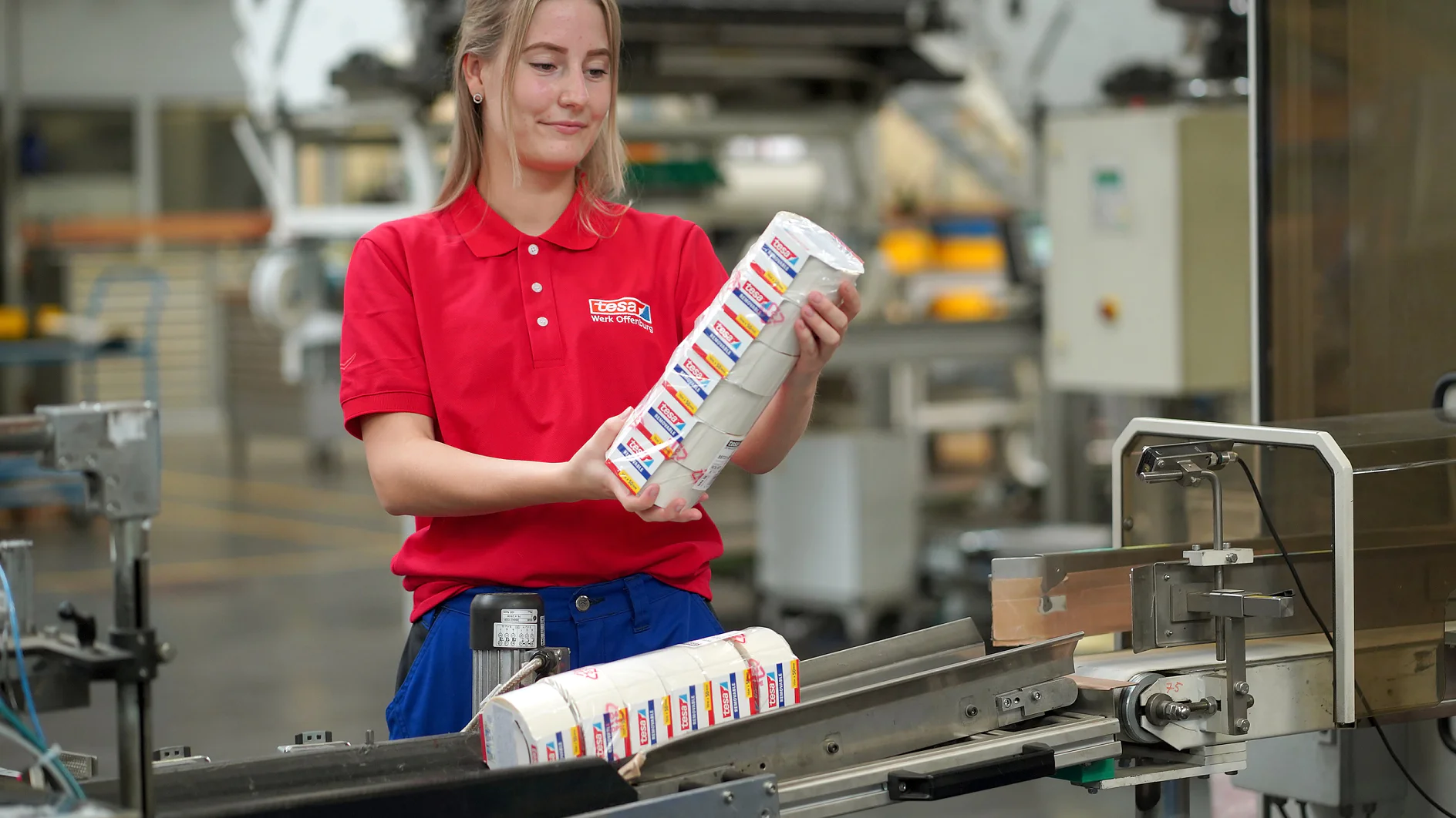 Eine Person in rotem Hemd und blauer Hose steht in einer Fabrik und legt verpackte Artikel auf ein Förderband. Im Hintergrund sind Industrieanlagen zu sehen, während des gesamten Vorgangs wird Tesa-Klebeband verwendet. (This text has been generated by AI)