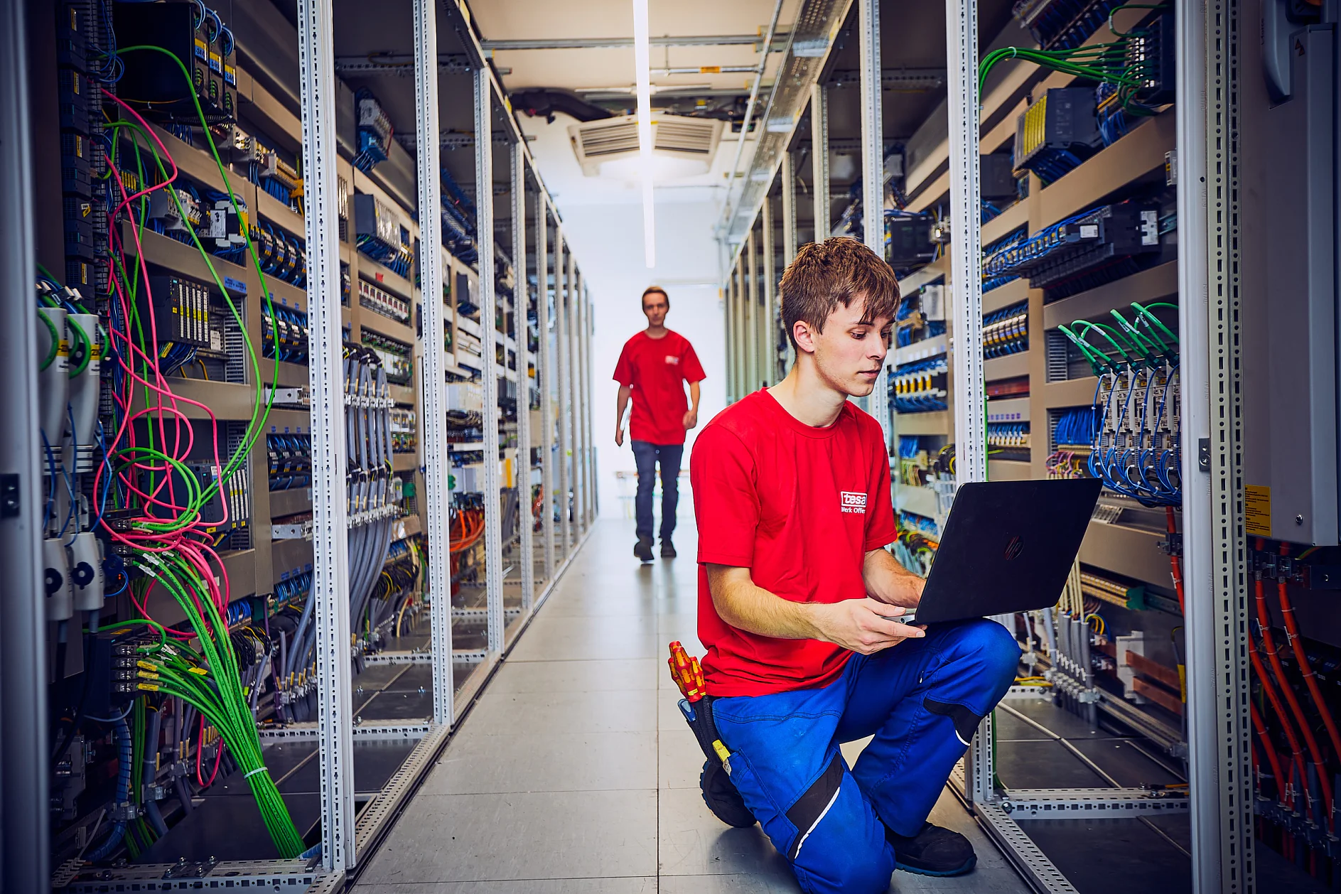 Zwei Personen in roten Hemden arbeiten in einem Rechenzentrum. Eine Person kniet mit einem Laptop neben Server-Racks, während die andere zwischen den Reihen der Geräte umhergeht. Der Raum ist voller Kabel, die mit Tesa-Klebeband befestigt und organisiert sind, und elektronischer Komponenten. (This text has been generated by AI)