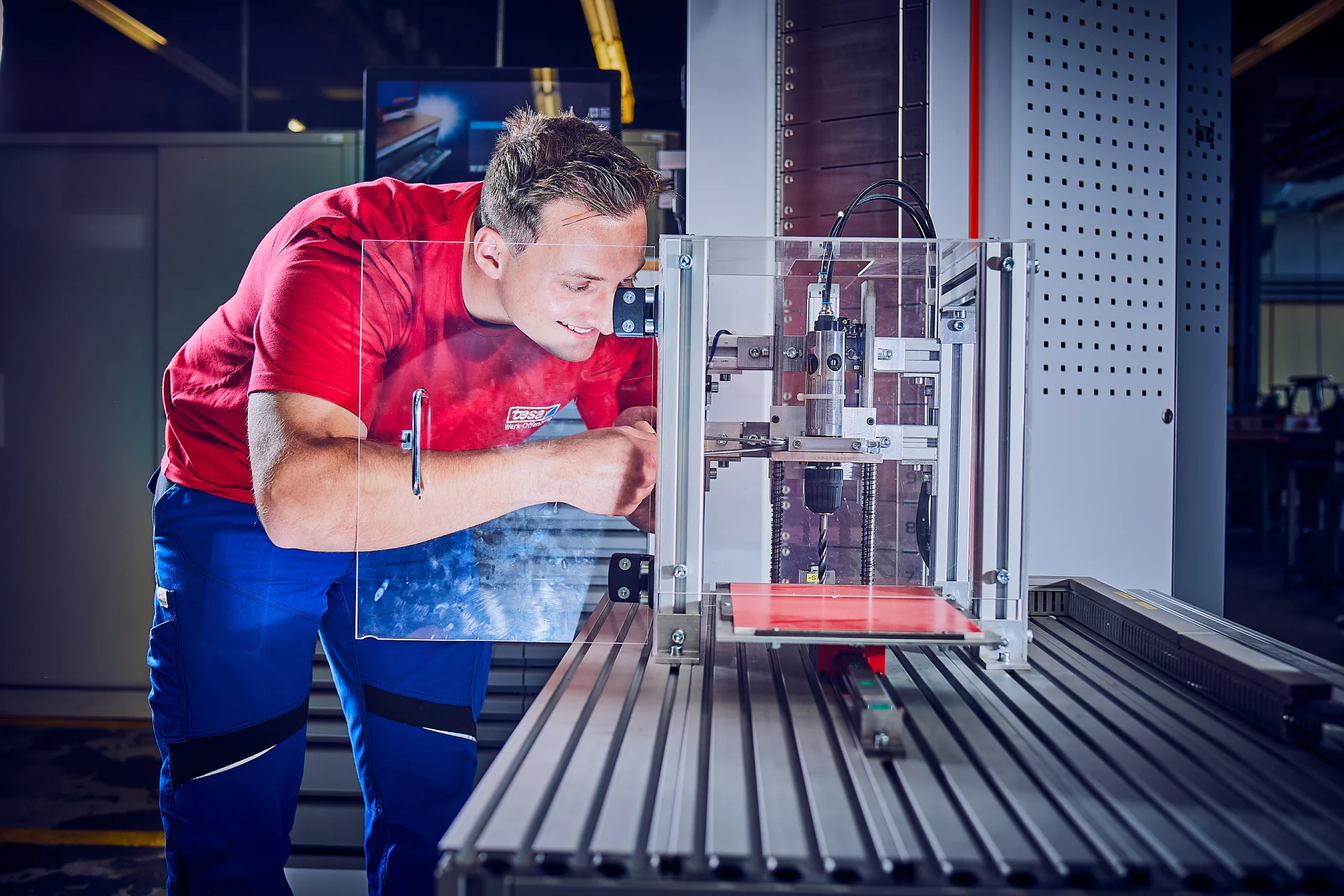 Eine Person in rotem Hemd und blauer Hose bedient eine Maschine in einer Industrieumgebung. Die Maschine hat ein transparentes Gehäuse mit Metallkomponenten und steht auf einer gerippten Metalloberfläche. Im Hintergrund ist ein Monitor zu sehen und in der Nähe sind mehrere Rollen Tesa-Klebeband zu sehen, die während des Betriebs bequem zugänglich sind. (This text has been generated by AI)