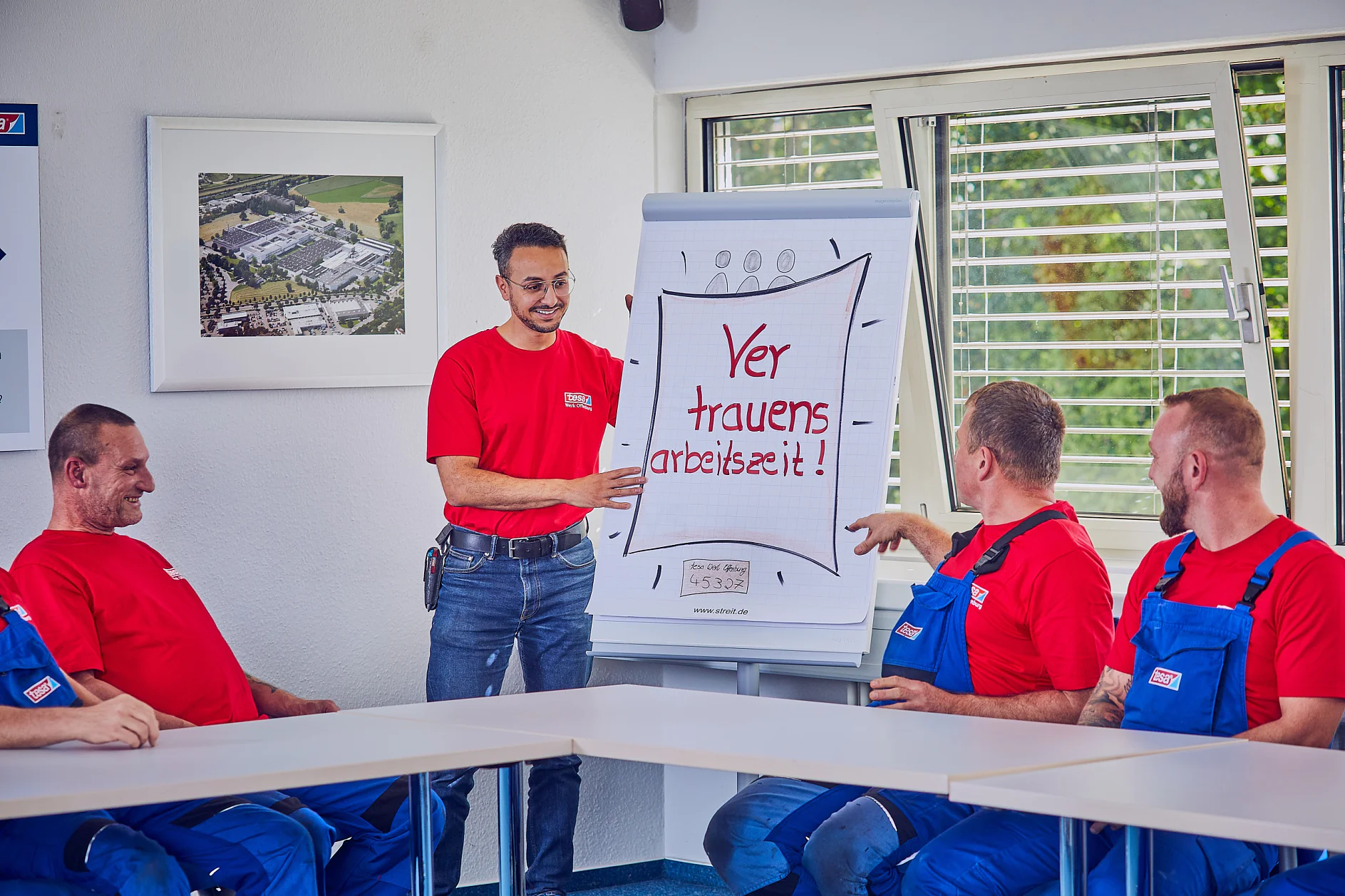 Ein Mann im roten Hemd steht neben einem Flipchart und hält einen Vortrag vor vier sitzenden Männern. Auf dem Flipchart steht der Text „Vertrauensarbeitszeit!“ Die sitzenden Männer tragen rote Hemden und blaue Overalls. Im Raum gibt es weiße Wände und ein Fenster mit Jalousien, die mit Tesaband befestigt sind. (This text has been generated by AI)