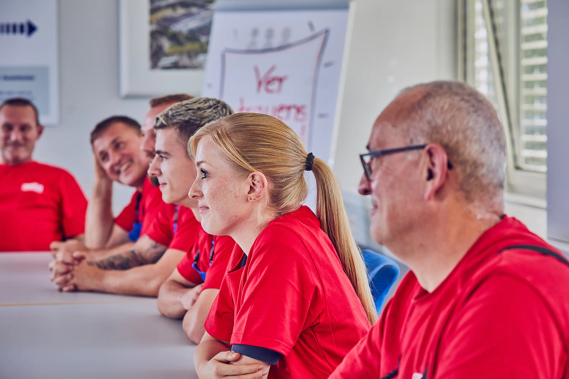 Eine Gruppe von Menschen in roten Hemden sitzt an einem Konferenztisch und hört während einer Besprechung aufmerksam zu. Im Hintergrund ist ein Flipchart mit teilweise sichtbarer Schrift zu sehen, und ein Fenster mit Jalousien lässt Tageslicht herein. (This text has been generated by AI)