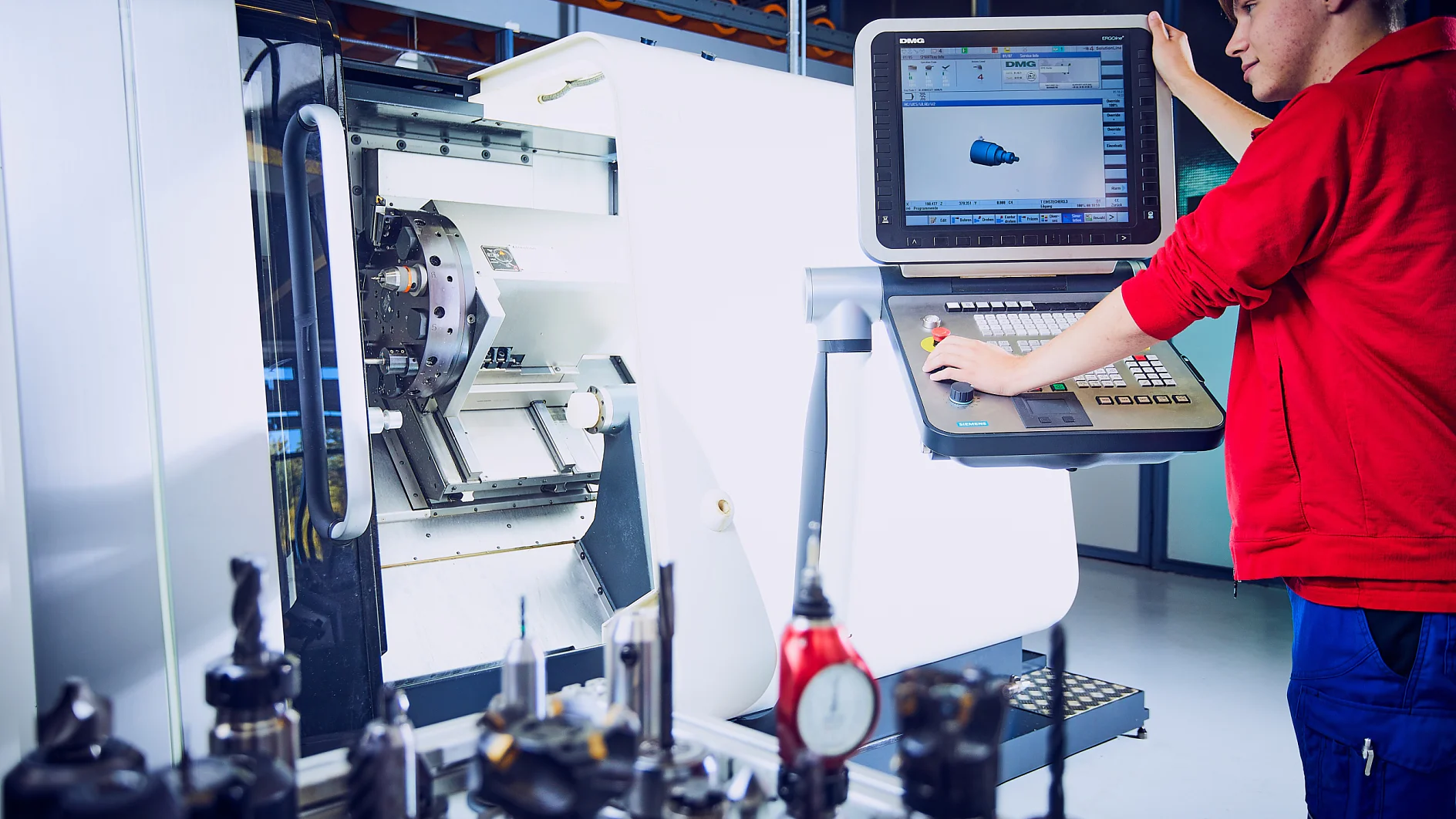 Ein Arbeiter im roten Hemd bedient eine CNC-Maschine und nimmt Einstellungen an einem großen Bedienfeld vor, auf dessen Bildschirm ein 3D-Modell angezeigt wird. Auf einer Oberfläche im Vordergrund sind verschiedene Werkzeugköpfe angeordnet, alle mit Tesa-Klebeband befestigt. (This text has been generated by AI)