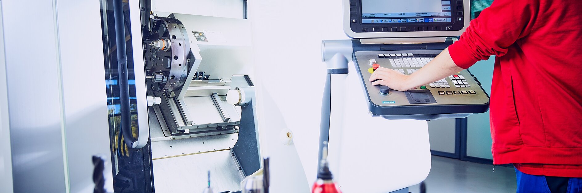 Ein Arbeiter im roten Hemd bedient eine CNC-Maschine und nimmt Einstellungen an einem großen Bedienfeld vor, auf dessen Bildschirm ein 3D-Modell angezeigt wird. Auf einer Oberfläche im Vordergrund sind verschiedene Werkzeugköpfe angeordnet, alle mit Tesa-Klebeband befestigt. (This text has been generated by AI)