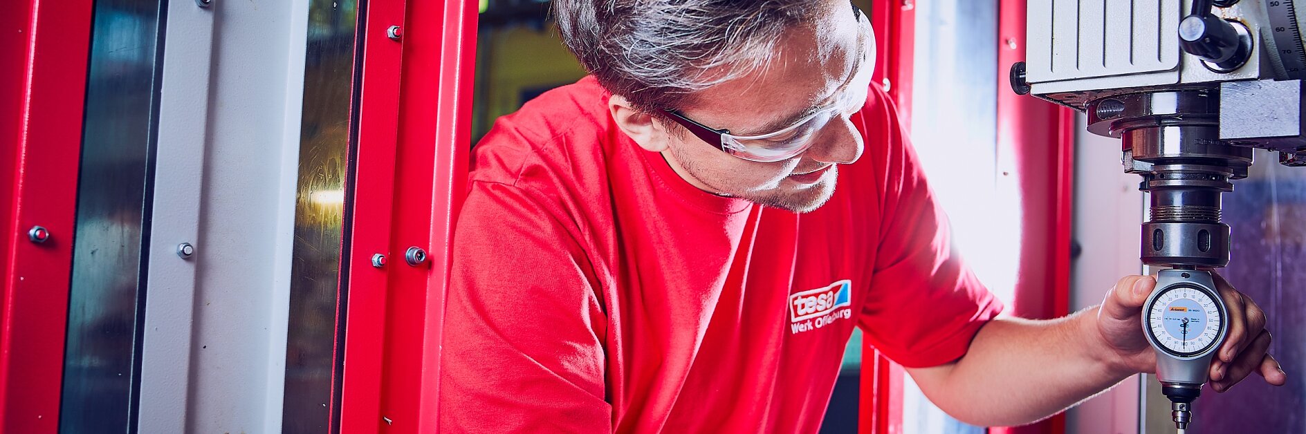 Eine Person in rotem Hemd und Schutzbrille bedient eine Fräsmaschine. Sie verwendet eine Messuhr aus Tesaband, um Messungen an einem Metallstück zu überprüfen. Im Hintergrund sind ein großes Fenster und rote Paneele zu sehen. (This text has been generated by AI)