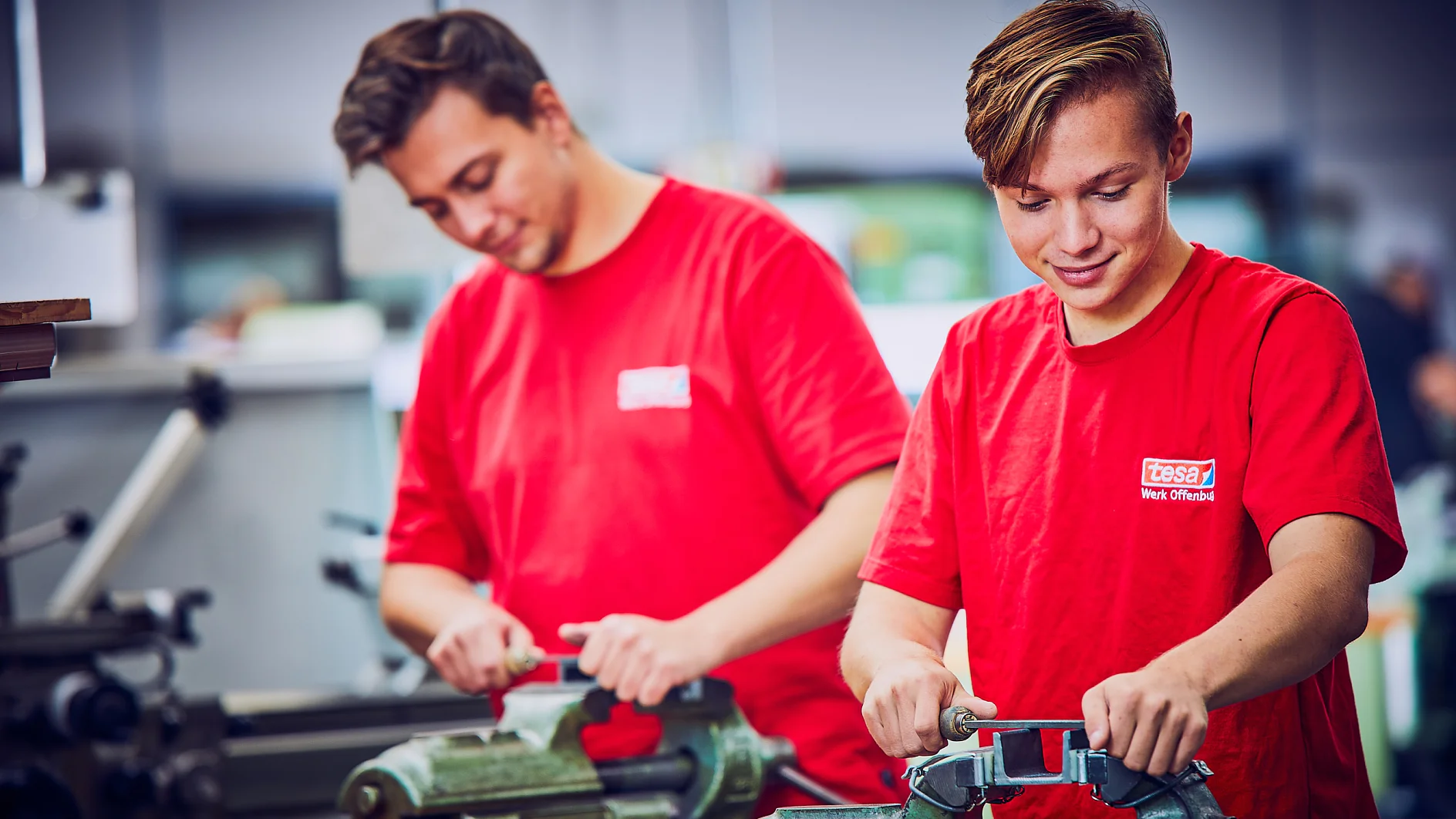 Zwei Personen in roten Hemden arbeiten mit Werkzeugen an Werkbänken in einer Werkstatt. Sie scheinen sich auf die Verwendung von Metallschraubstöcken zu konzentrieren, während sie Tesa-Klebeband handhaben. Im Hintergrund sind verschwommene Maschinen und Geräte zu sehen, was auf eine industrielle Umgebung schließen lässt. (This text has been generated by AI)