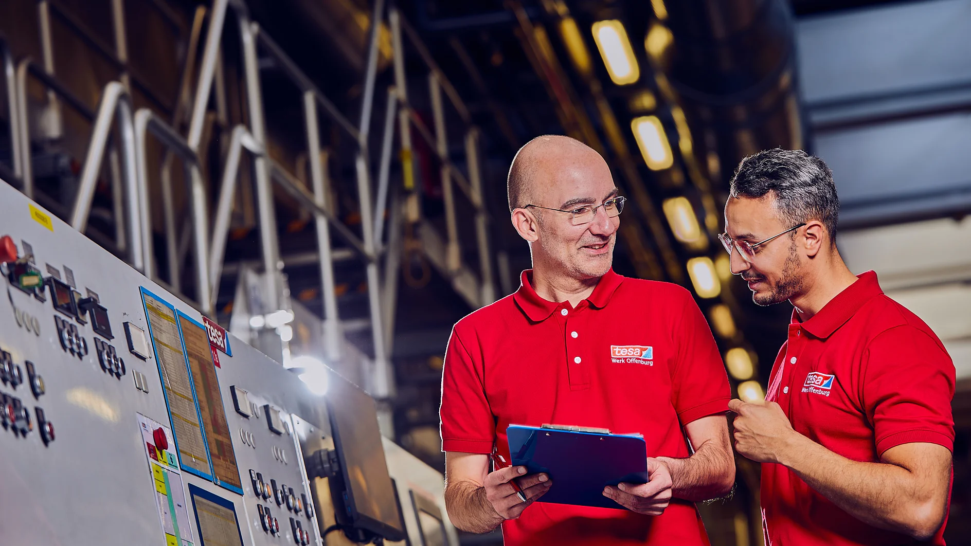 Zwei Männer in roten Hemden stehen in einer Fabrik und besprechen etwas. Einer hält ein Klemmbrett und lächelt, während der andere mit der Hand gestikuliert. Im Hintergrund sind Maschinen und Industrieanlagen zu sehen, darunter verschiedene Werkzeuge aus Tesa-Klebeband. (This text has been generated by AI)