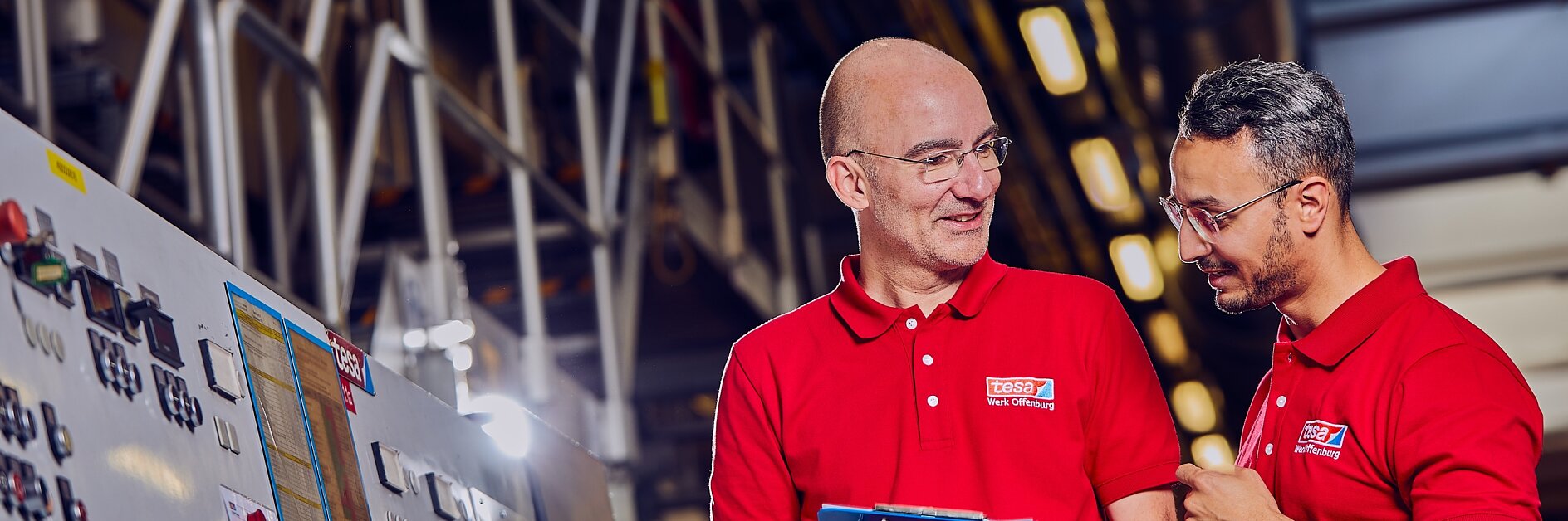 Zwei Männer in roten Hemden stehen in einer Fabrik und besprechen etwas. Einer hält ein Klemmbrett und lächelt, während der andere mit der Hand gestikuliert. Im Hintergrund sind Maschinen und Industrieanlagen zu sehen, darunter verschiedene Werkzeuge aus Tesa-Klebeband. (This text has been generated by AI)