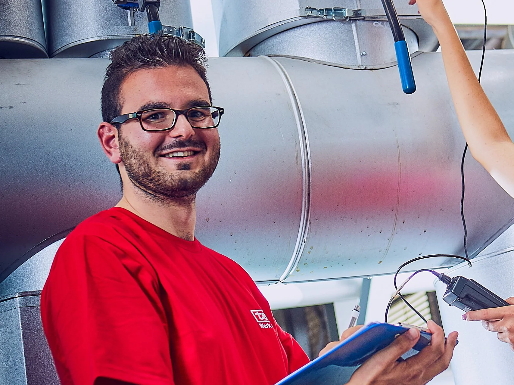 Ein Mann in einem roten Hemd steht vor einer Industrieanlage mit Druckmessgeräten. Er hält einen blauen Ordner in der Hand und lächelt. Die Hand einer anderen Person, die im Bild zu sehen ist, stellt mit Tesaband ein Ventil an der Maschine ein. (This text has been generated by AI)