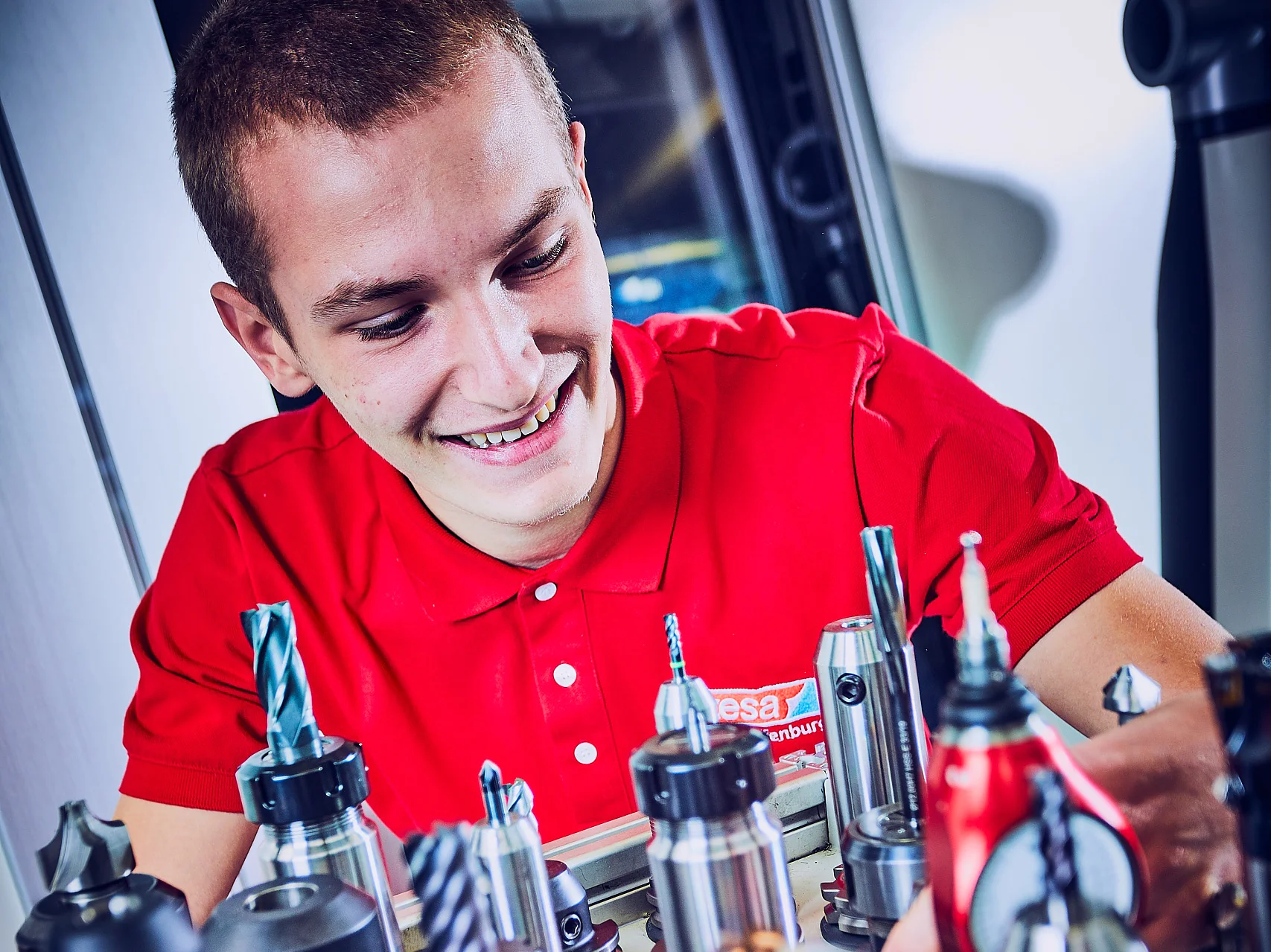 Eine Person in einem roten Hemd lächelt und richtet mehrere Metallwerkzeuge, die aufrecht auf einem Gestell angeordnet sind, mit Tesa-Klebeband aus. Die Werkzeuge haben verschiedene Formen und Größen und werden wahrscheinlich in der maschinellen Bearbeitung oder Fertigung verwendet. Die Umgebung scheint eine Werkstatt oder ein Industrieumfeld zu sein. (This text has been generated by AI)