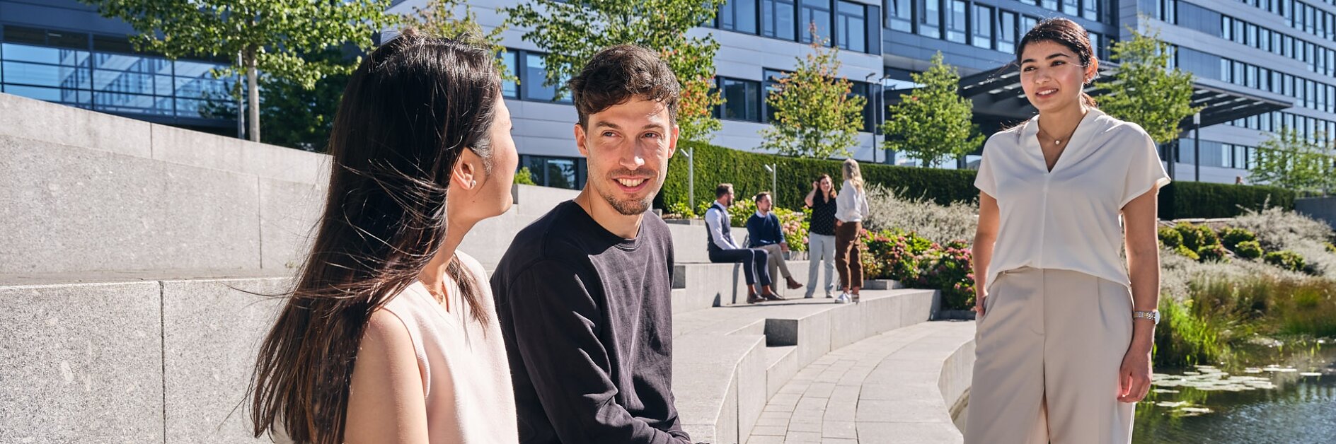 Drei Personen unterhalten sich im Freien in der Nähe eines Teichs und eines modernen Bürogebäudes. Zwei sitzen auf Steinstufen, während einer steht. Bäume und Pflanzen umgeben den Bereich, und im Hintergrund sind weitere Personen zu sehen. (This text has been generated by AI)