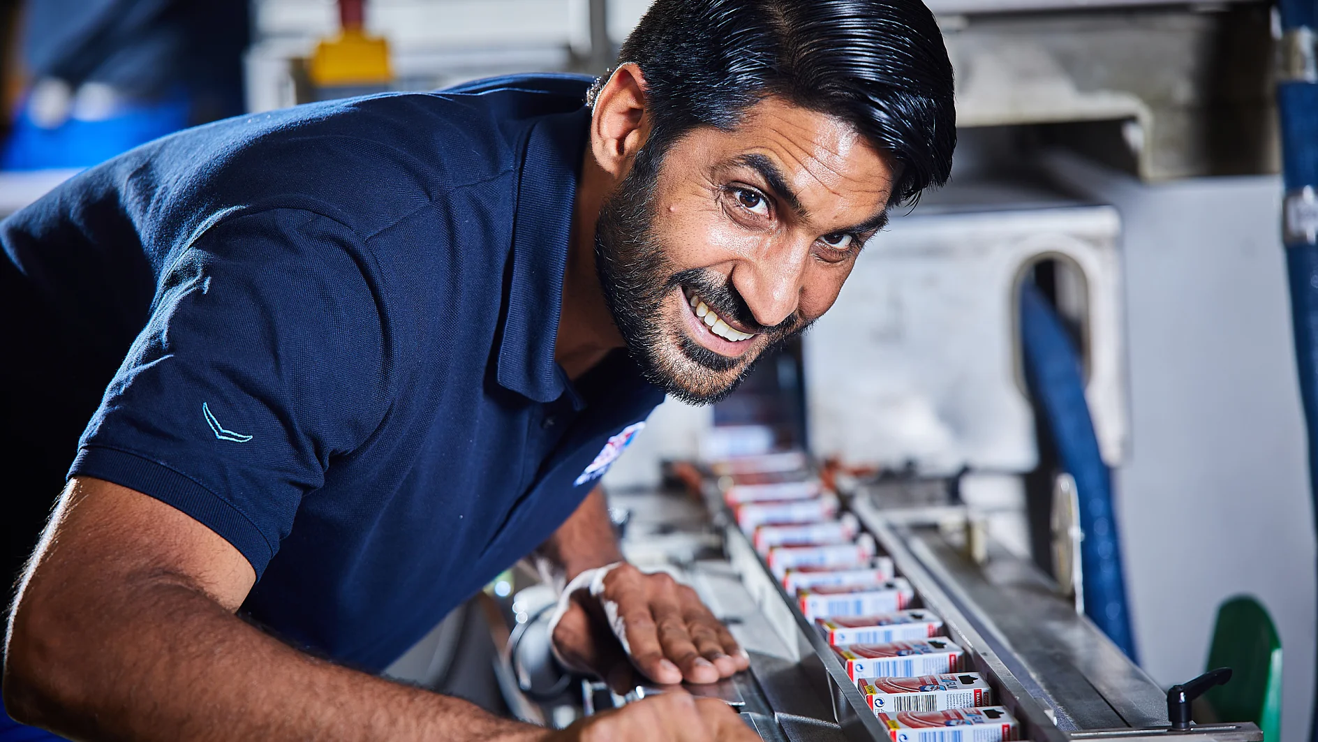 Ein Mann in einem marineblauen Poloshirt beugt sich über eine Maschine, um sie zu inspizieren oder daran zu arbeiten. Mehrere kleine Kästen sind auf der Oberfläche der Maschine aufgereiht. Die Umgebung scheint industriell oder in einer Werkstatt zu sein. Der Mann blickt lächelnd in die Kamera. (This text has been generated by AI)