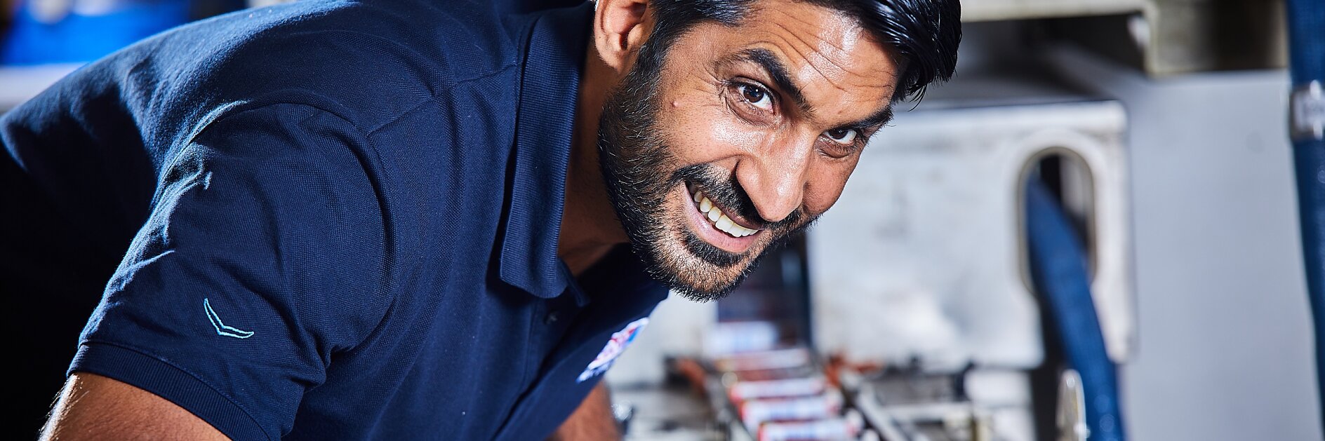 Ein Mann in einem marineblauen Poloshirt beugt sich über eine Maschine, um sie zu inspizieren oder daran zu arbeiten. Mehrere kleine Kästen sind auf der Oberfläche der Maschine aufgereiht. Die Umgebung scheint industriell oder in einer Werkstatt zu sein. Der Mann blickt lächelnd in die Kamera. (This text has been generated by AI)