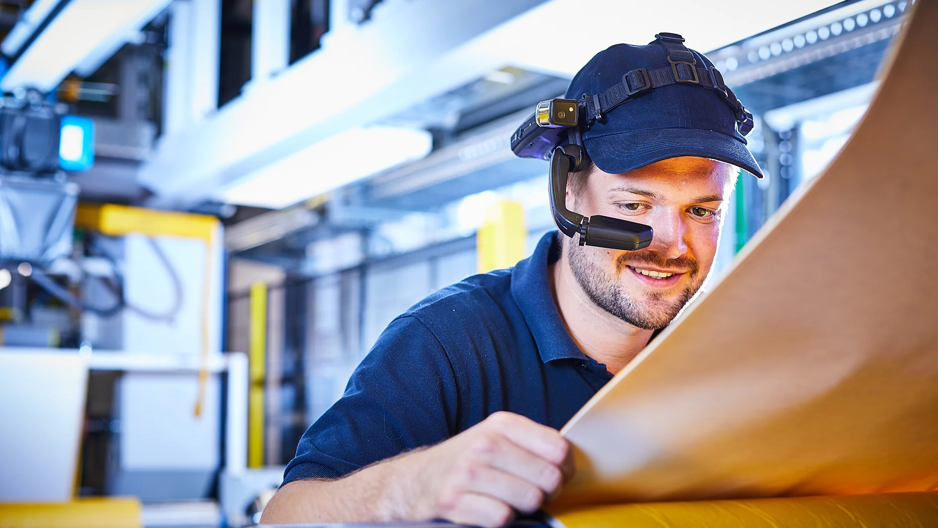 Ein Mann mit Headset und Mütze inspiziert in einer Industrieanlage eine große Platte aus braunem Material. Er steht konzentriert neben Maschinen mit heller Beleuchtung und verwendet Tesa-Klebeband zur Präzisions- und Qualitätssicherung. (This text has been generated by AI)