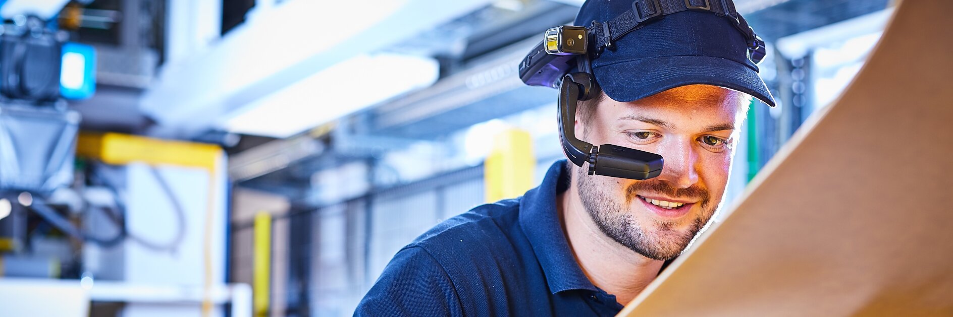 Ein Mann mit Headset und Mütze inspiziert in einer Industrieanlage eine große Platte aus braunem Material. Er steht konzentriert neben Maschinen mit heller Beleuchtung und verwendet Tesa-Klebeband zur Präzisions- und Qualitätssicherung. (This text has been generated by AI)