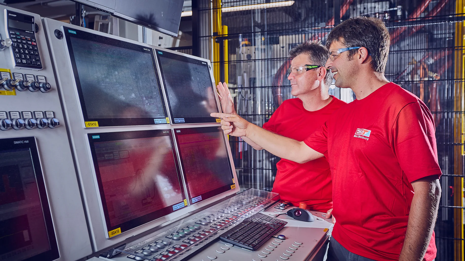 Zwei Männer in roten Hemden und Schutzbrillen blicken auf eine Reihe von vier Computerbildschirmen, die auf einem Bedienfeld montiert sind, und zeigen darauf. Die Szene scheint eine Industrie- oder Fabrikumgebung mit Maschinen im Hintergrund zu sein. (This text has been generated by AI)