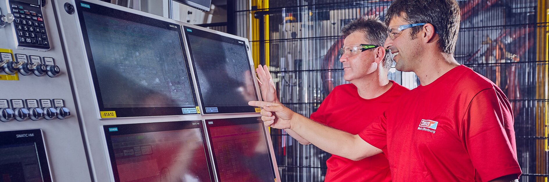 Zwei Männer in roten Hemden und Schutzbrillen blicken auf eine Reihe von vier Computerbildschirmen, die auf einem Bedienfeld montiert sind, und zeigen darauf. Die Szene scheint eine Industrie- oder Fabrikumgebung mit Maschinen im Hintergrund zu sein. (This text has been generated by AI)