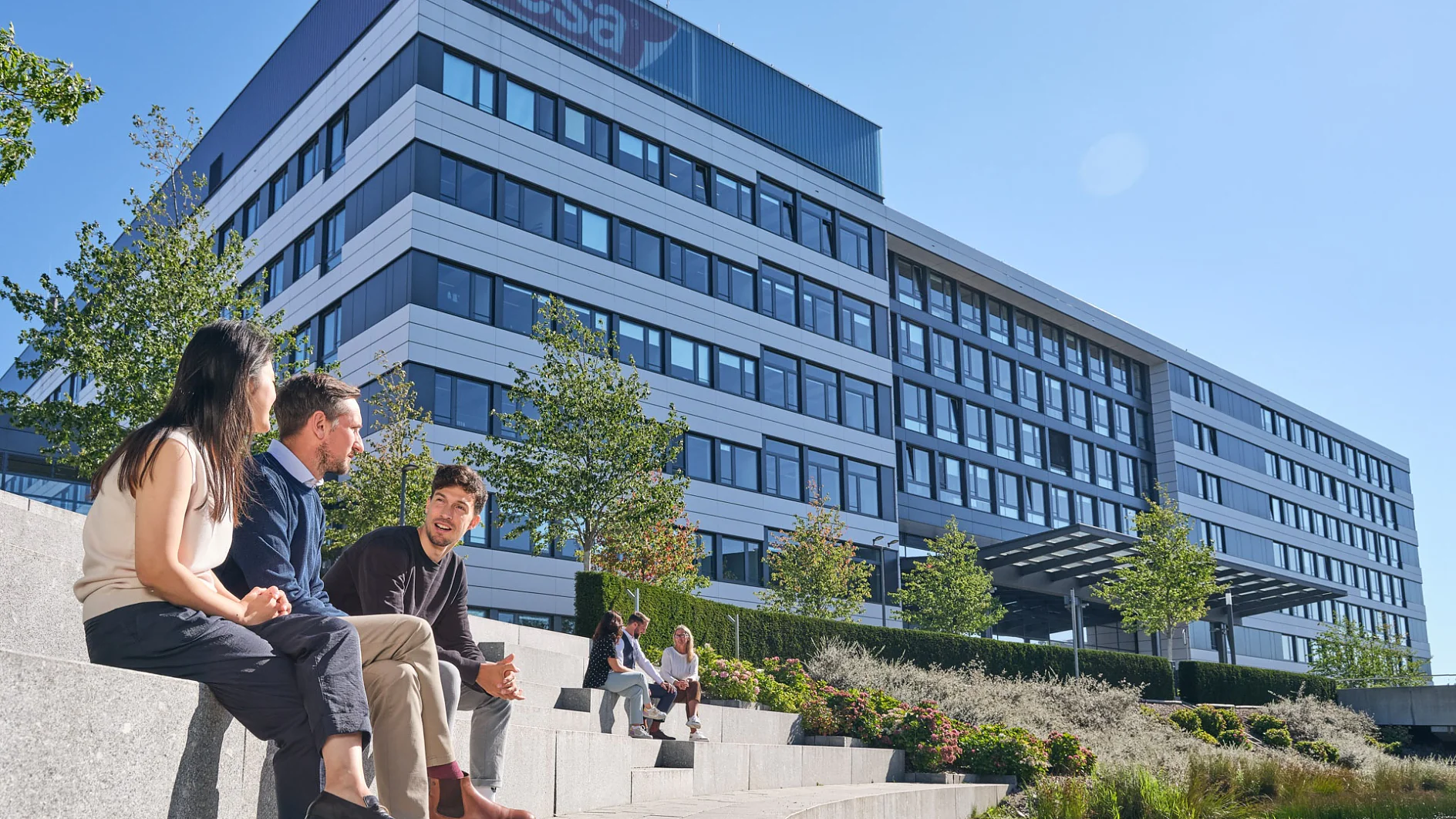 Drei Personen sitzen auf Steinstufen neben einem Teich vor einem modernen Bürogebäude mit einem „Tesa Tape“-Logo. Das Gebäude ist mehrstöckig und hat große Fenster. Es gibt Bäume und Landschaftsgestaltung und der Himmel ist klar. (This text has been generated by AI)
