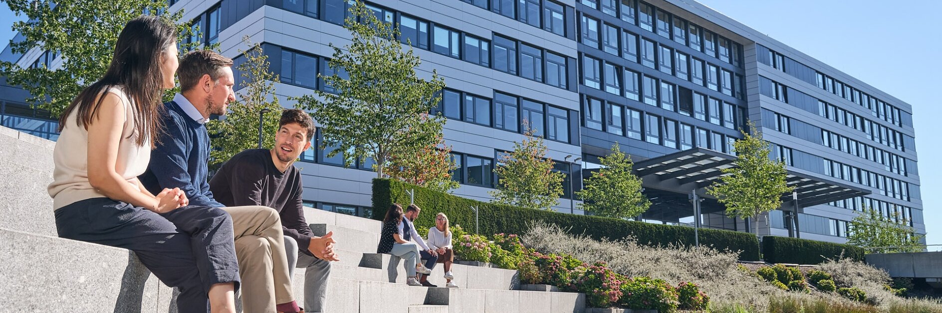 Drei Personen sitzen auf Steinstufen neben einem Teich vor einem modernen Bürogebäude mit einem „Tesa Tape“-Logo. Das Gebäude ist mehrstöckig und hat große Fenster. Es gibt Bäume und Landschaftsgestaltung und der Himmel ist klar. (This text has been generated by AI)