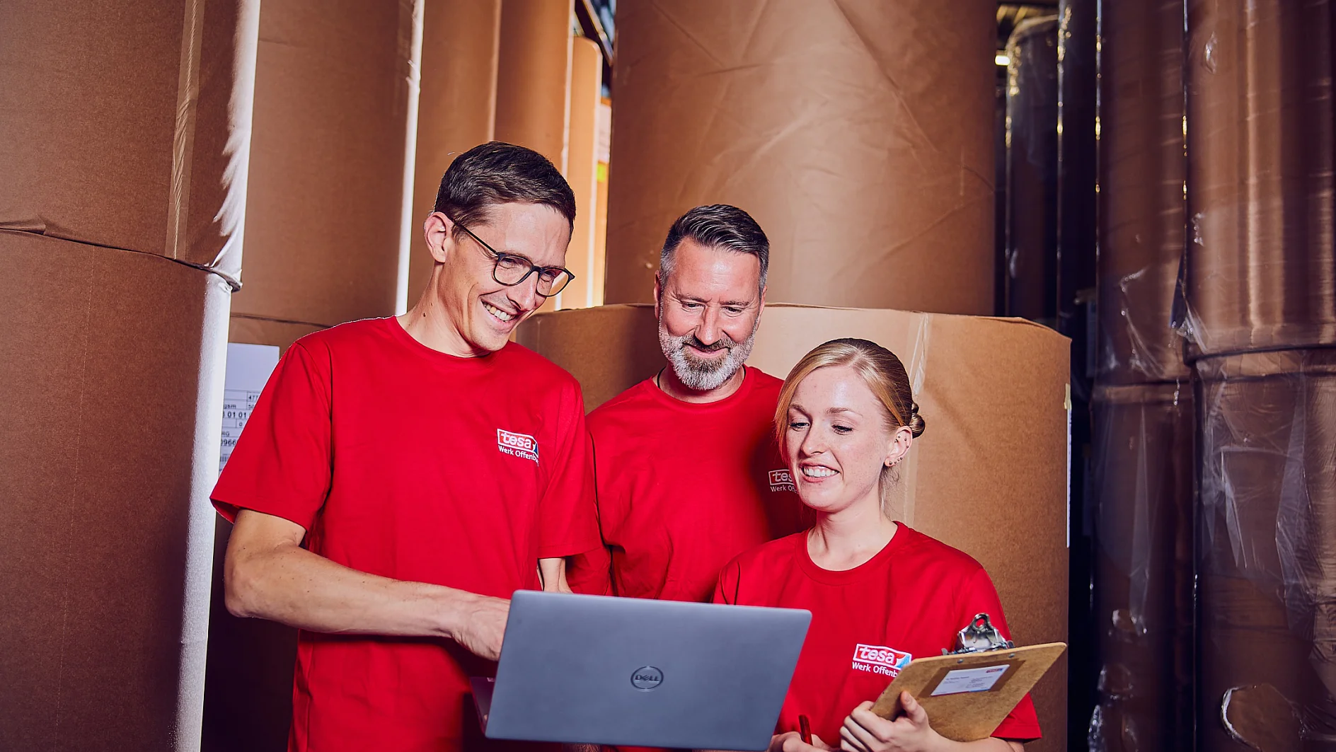 Drei Personen in roten Hemden stehen zwischen großen Rollen braunen Papiers. Eine Person hält einen Laptop, eine andere ein Klemmbrett, und sie scheinen Informationen zu besprechen oder zu überprüfen. Die Umgebung ähnelt einem Lagerhaus oder einer Lagerhalle. (This text has been generated by AI)