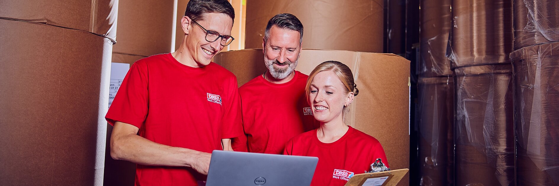 Drei Personen in roten Hemden stehen zwischen großen Rollen braunen Papiers. Eine Person hält einen Laptop, eine andere ein Klemmbrett, und sie scheinen Informationen zu besprechen oder zu überprüfen. Die Umgebung ähnelt einem Lagerhaus oder einer Lagerhalle. (This text has been generated by AI)