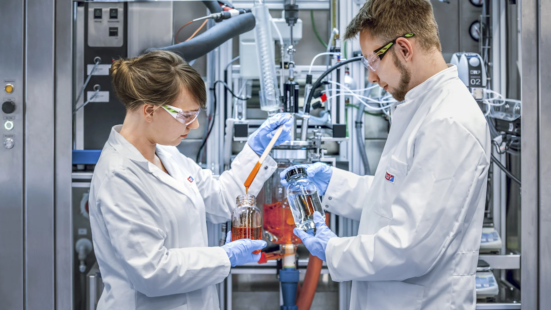 Zwei Wissenschaftler in weißen Laborkitteln und Schutzbrillen arbeiten in einem Industrielabor mit Flüssigkeitsproben in Gefäßen. Sie stehen vor komplexen Maschinen und konzentrieren sich auf ihre Aufgaben. Im Hintergrund sind Rohre und Tesa-Klebeband-Geräte zu sehen. (This text has been generated by AI)