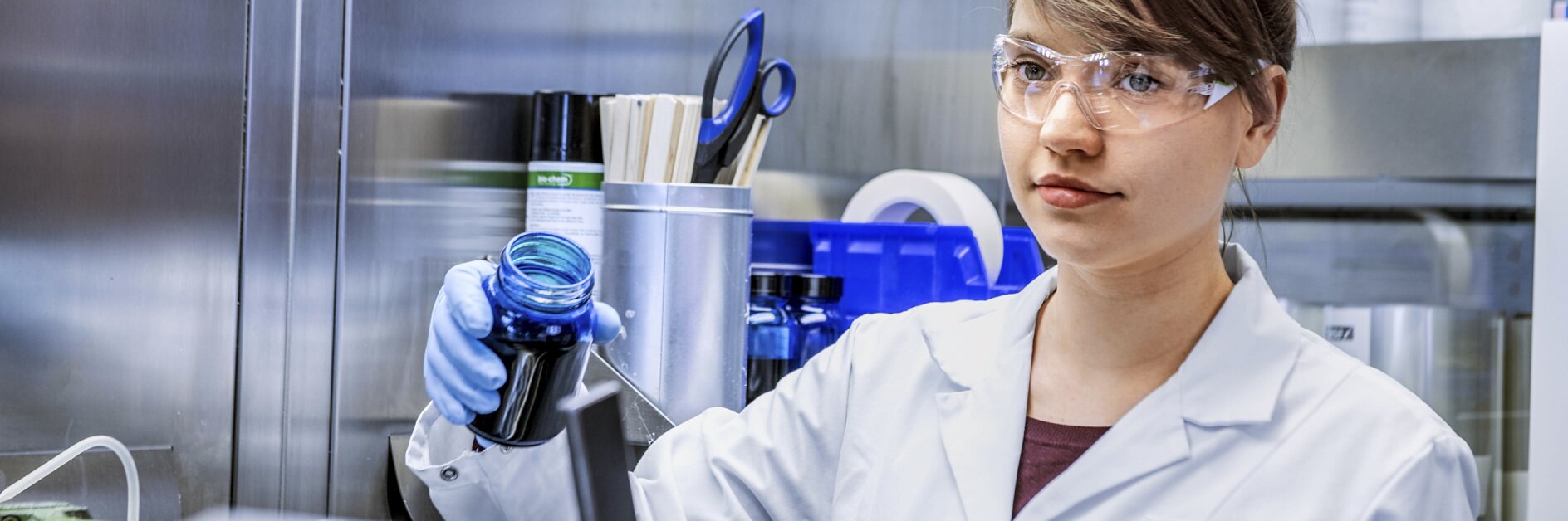 Eine Person im Laborkittel und mit Schutzbrille hält in einer Laborumgebung ein Gefäß mit blauer Flüssigkeit. Sie ist von Laborgeräten, Lagerregalen und Tesa-Klebeband umgeben. (This text has been generated by AI)