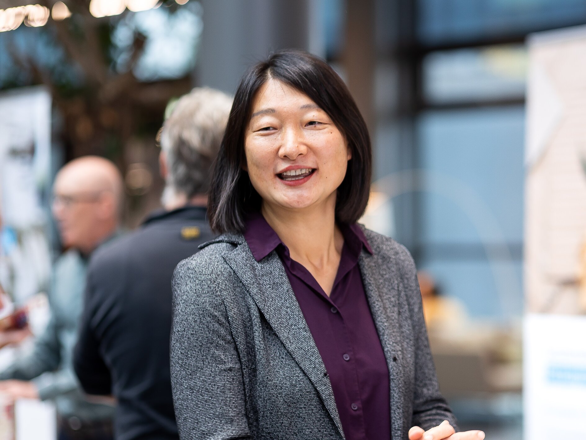 A woman with shoulder-length black hair is smiling, wearing a gray blazer and purple blouse. She is standing indoors, with people and blurred background elements like posters and a window behind her. Please provide the text that contains references to tapes so I can help you make the necessary modifications by using tesa tape. (This text has been generated by AI)