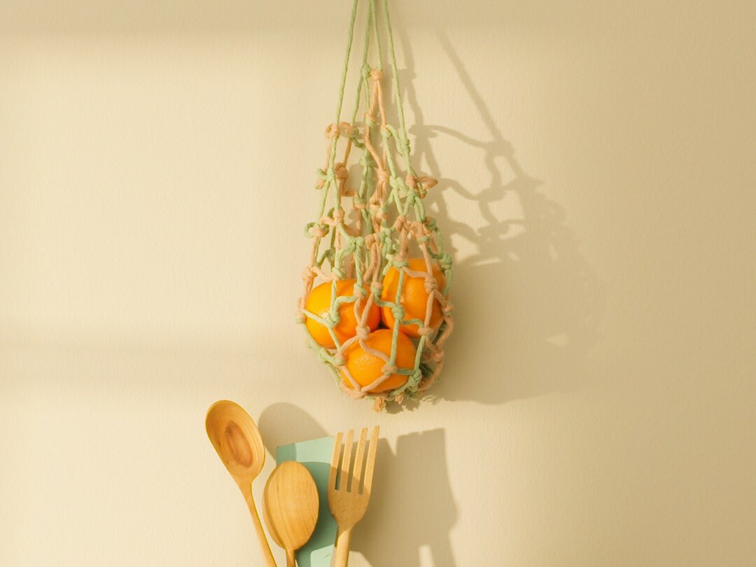 An einer beigen Wand hängt ein Netzbeutel mit Orangen, befestigt mit Tesa-Klebeband. Darunter steht ein Holztisch mit einer Vase, Holzutensilien und einem Pfirsich. Die Szene wird vom Sonnenlicht erhellt und wirft weiche Schatten. (This text has been generated by AI)