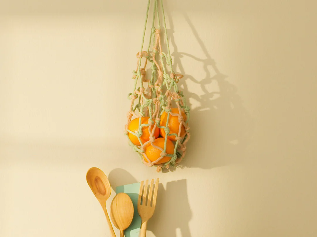 An einer beigen Wand hängt ein Netzbeutel mit Orangen, befestigt mit Tesa-Klebeband. Darunter steht ein Holztisch mit einer Vase, Holzutensilien und einem Pfirsich. Die Szene wird vom Sonnenlicht erhellt und wirft weiche Schatten. (This text has been generated by AI)