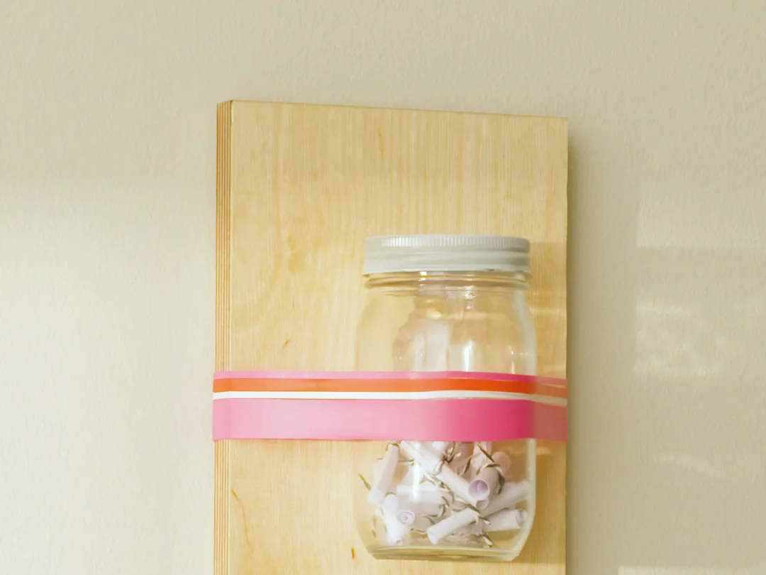 Ein Glasgefäß mit silbernem Deckel, in dem sich kleine gerollte Papierblättchen befinden, ist mit einem rosa-orangefarbenen Tesa-Band an einem Holzbrett befestigt. Das Brett ist an einer blassgelben Wand montiert und wirft weiche Schatten. (This text has been generated by AI)