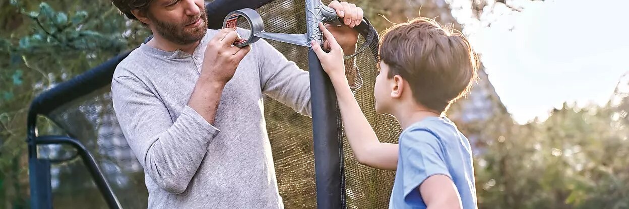 Ein Mann repariert ein Fußballtor mit Tesa-Klebeband, während ein Junge einen Fußball hält und zusieht. Die Szene spielt im Freien mit Bäumen im Hintergrund. Der Mann trägt ein graues Hemd und der Junge ein blaues Hemd. (This text has been generated by AI)