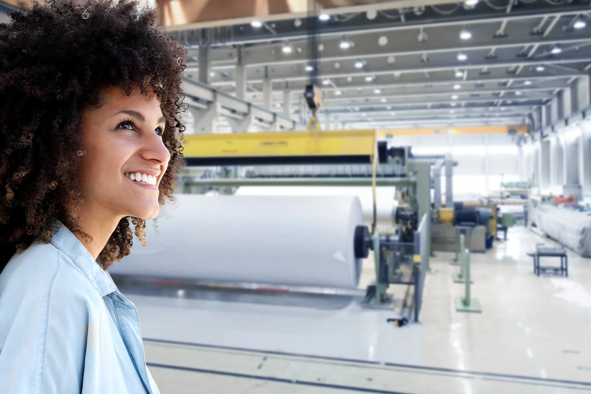 A woman with curly hair smiles while standing in a spacious industrial facility. Behind her, a large machine processes a substantial roll of white tesa tape in a well-lit, modern factory setting. (This text has been generated by AI)