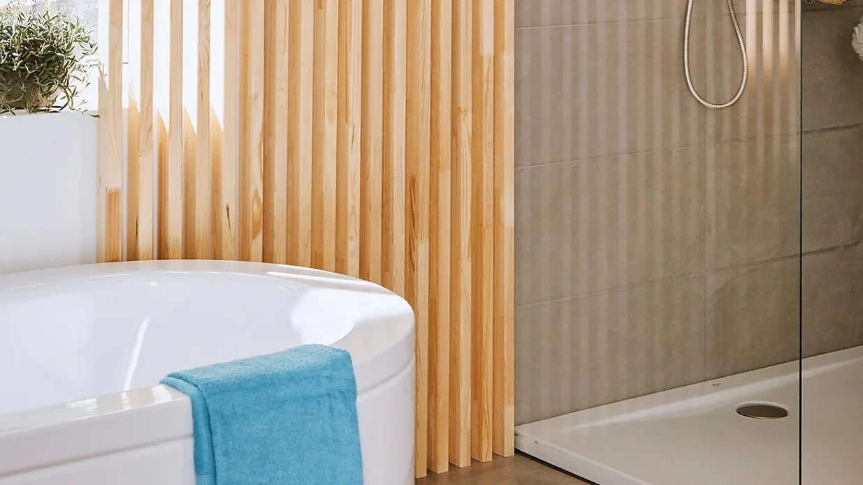 badezimmerdekorieren_1280x1280 Ein modernes Badezimmer verfügt über eine weiße Eckbadewanne mit einem über den Rand hängenden blauen Handtuch, Wandpaneele mit Holzlatten, eine Topfpflanze und eine ebenerdige Dusche mit beigen Fliesen und einer Glastrennwand. (This text has been generated by AI)