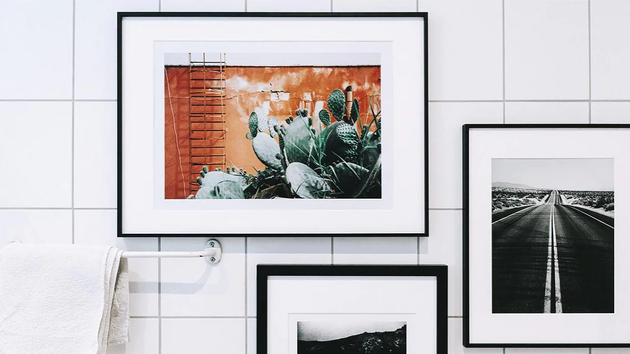 badezimmerdekorieren-tipps-wanddekoration_1280x1280 An einer gefliesten Wand hängen gerahmte Kunstwerke, darunter ein farbenfrohes Gemälde von Kakteen auf orangefarbenem Hintergrund und zwei Schwarzweißfotos. An einer Stange in der Nähe hängt ein weißes Handtuch, das mit Tesa-Klebeband befestigt ist. (This text has been generated by AI)