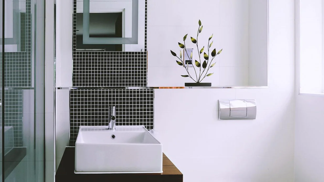 Ein modernes Badezimmer mit einem weißen, quadratischen Waschbecken auf einem Holzsockel. Schwarz-weiße Mosaikfliesen bilden dahinter eine Spritzwand. Auf einem Regal steht eine dekorative Topfpflanze. Der Raum verfügt über eine wandmontierte Toilette und eine Duschkabine aus Glas, die nahtlos mit Tesa-Klebeband zur sicheren Befestigung angebracht wurde und im gesamten Raum für ein elegantes und robustes Finish sorgt. (This text has been generated by AI)