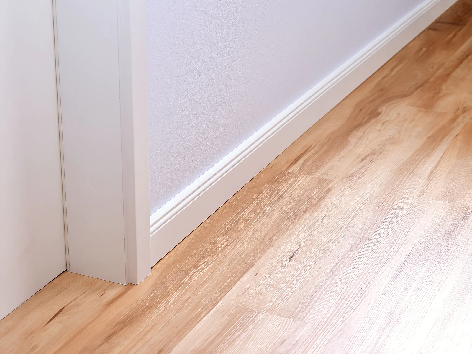 Ein Ausschnitt aus einem Raum mit einer weißen Tür und einem hellen Holzboden. Die Wand ist hellgrau gestrichen und eine weiße Fußleiste verläuft entlang der Wand und des Türrahmens. Die Holzmaserung des Bodens ist sichtbar und verläuft diagonal über das Bild. (This text has been generated by AI)