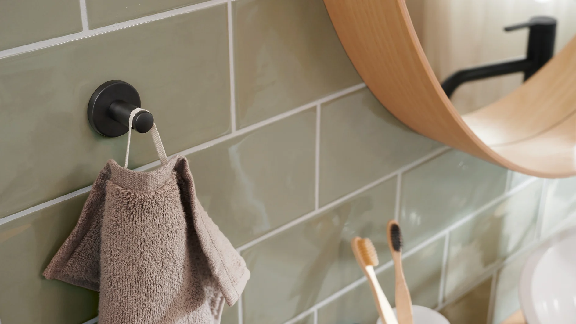 Eine Badezimmerszene mit einem grauen Handtuch, das an einem Haken an einer Wand aus grünen Fliesen hängt. Auf der rechten Seite ist teilweise ein Spiegel mit Holzrahmen sichtbar und zwei Zahnbürsten liegen in einem Halter auf einer weißen Arbeitsplatte. (This text has been generated by AI)