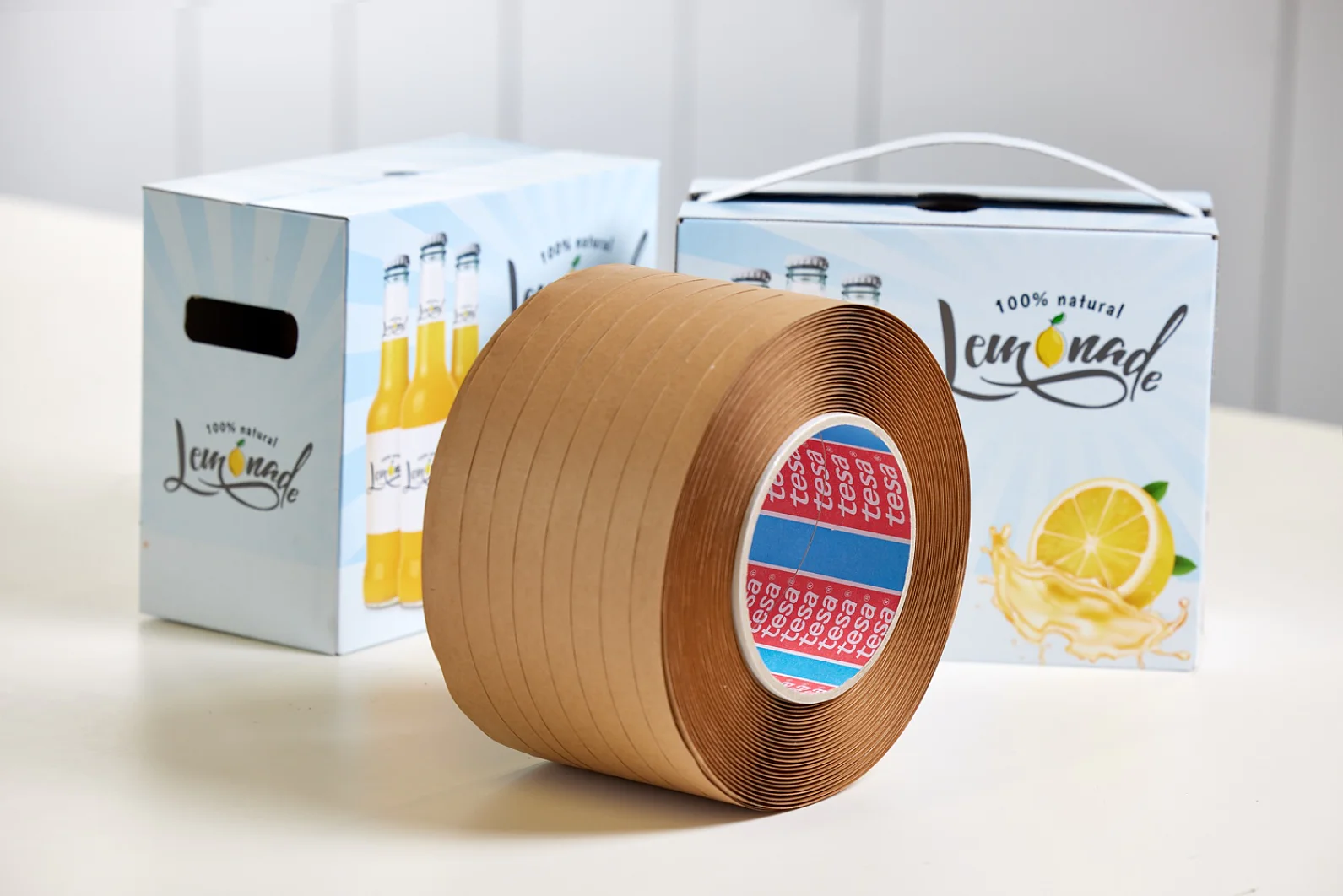 A roll of brown tesa tape is on a white surface. In the background, there are two boxes labeled 100% natural lemonade, with lemon graphics. The boxes have a handle and an illustration of lemonade bottles. (This text has been generated by AI)