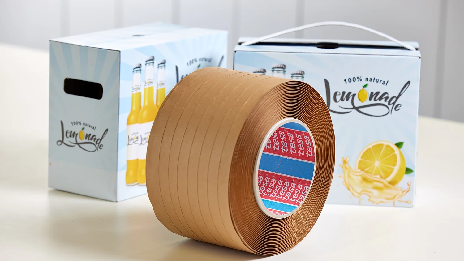 A roll of brown tesa tape is on a white surface. In the background, there are two boxes labeled 100% natural lemonade, with lemon graphics. The boxes have a handle and an illustration of lemonade bottles. (This text has been generated by AI)