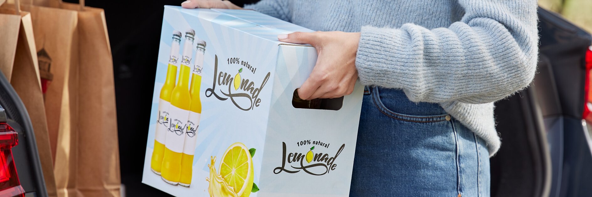 A person wearing a gray sweater and blue jeans is loading a box labeled Lemonade into the trunk of a car. The trunk also contains brown paper bags sealed with tesa tape.