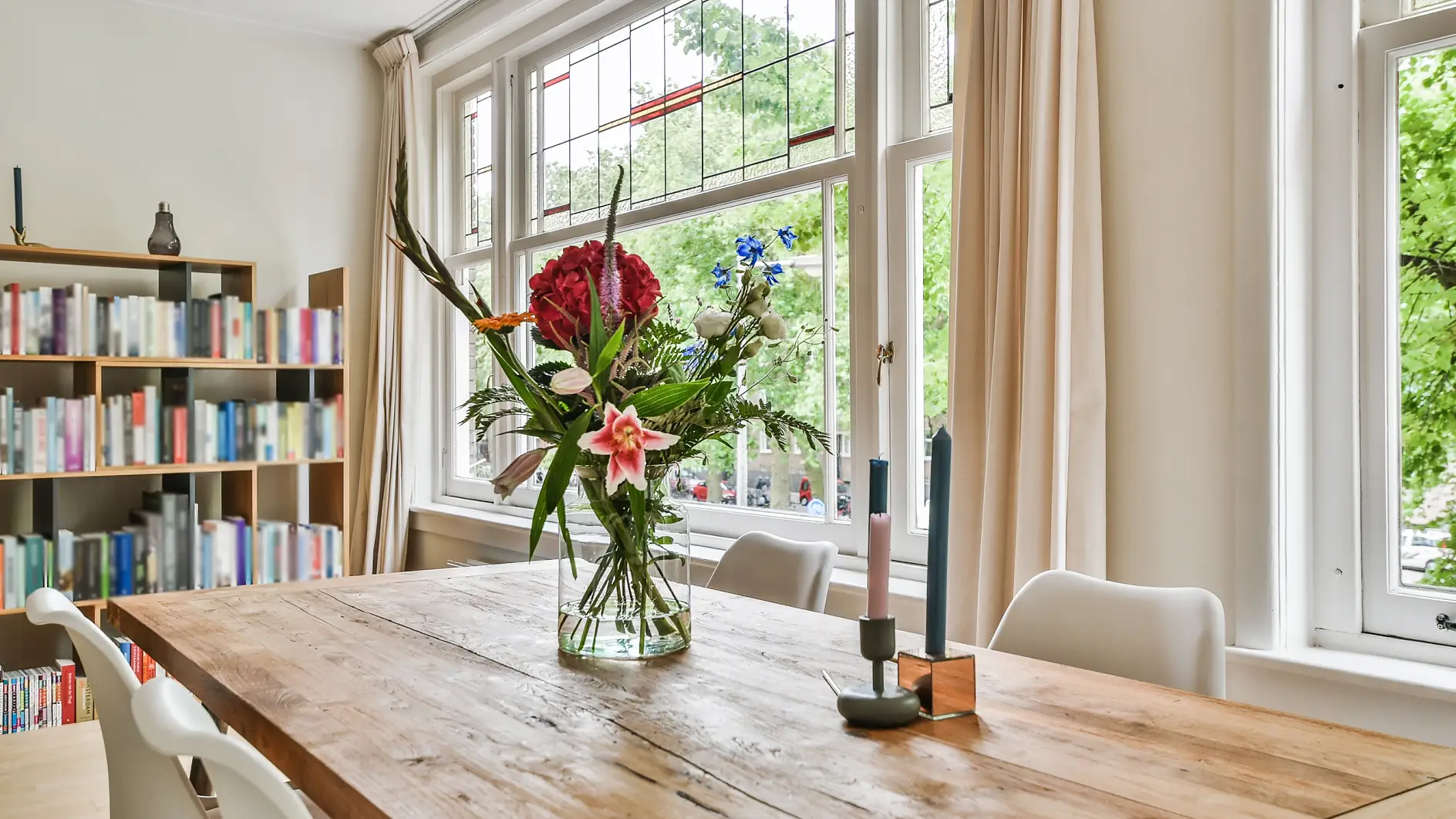 Glass vase with fresh flowers and stylish candles placed on wooden table against windows and bookcase in light dining room